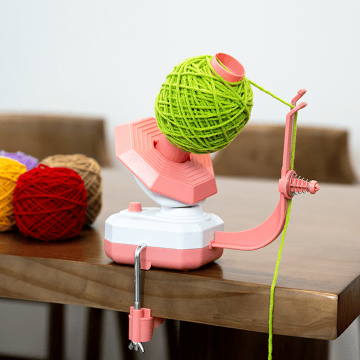 An Electric Yarn Winder in pink and white is clamped to a wooden table, winding a green yarn ball, with other colorful yarn balls visible in the background.