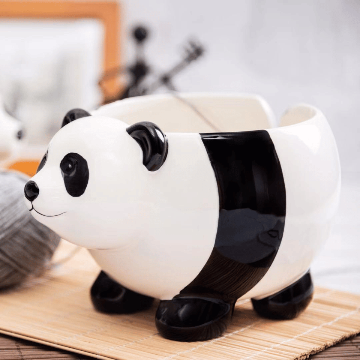 The Panda Ceramic Yarn Bowl sits on a bamboo mat with a gray yarn ball nearby—ideal for beginner-friendly projects like cozy blankets—against a blurred background.