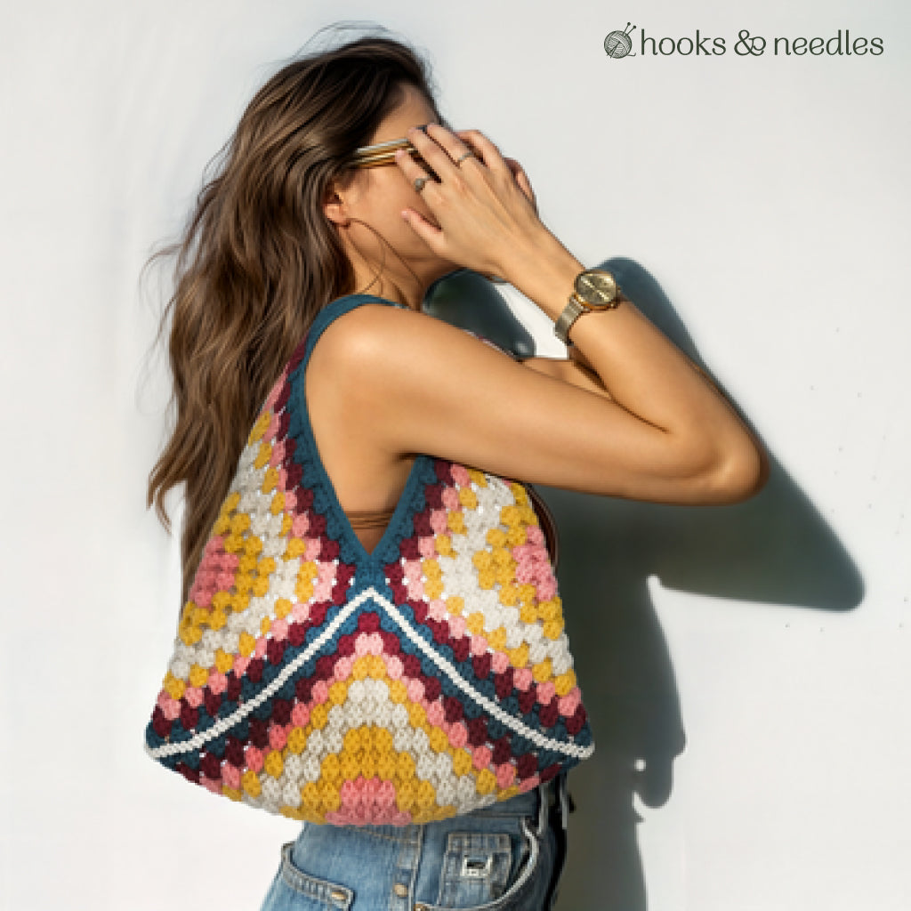 A woman in sunglasses carries a vibrant tote made from the Granny Square Tote Bag Crochet Pattern. The "hooks & needles" logo appears in the top right. Ideal for anyone seeking a stylish, beginner-friendly crochet project.