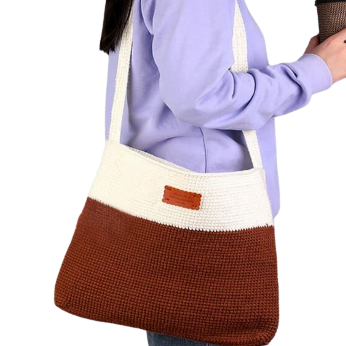 A person in a lavender sweater carries the Modern Crochet Tote Bag Kit, crafted from white and brown cotton yarn, while holding a coffee cup.