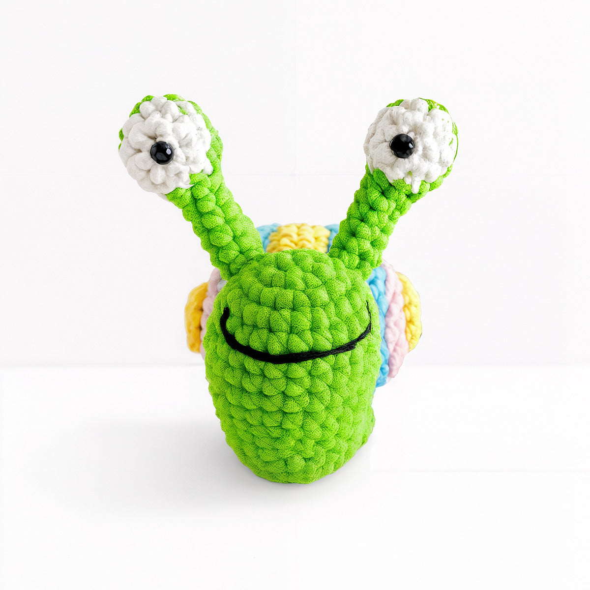 Speedy The Snail Crochet Pattern features a green amigurumi snail with big white-and-black eyes on stalks, a black smile, and a yellow, pink, and blue shell. Ideal for anyone wanting an adorable crochet snail project.
