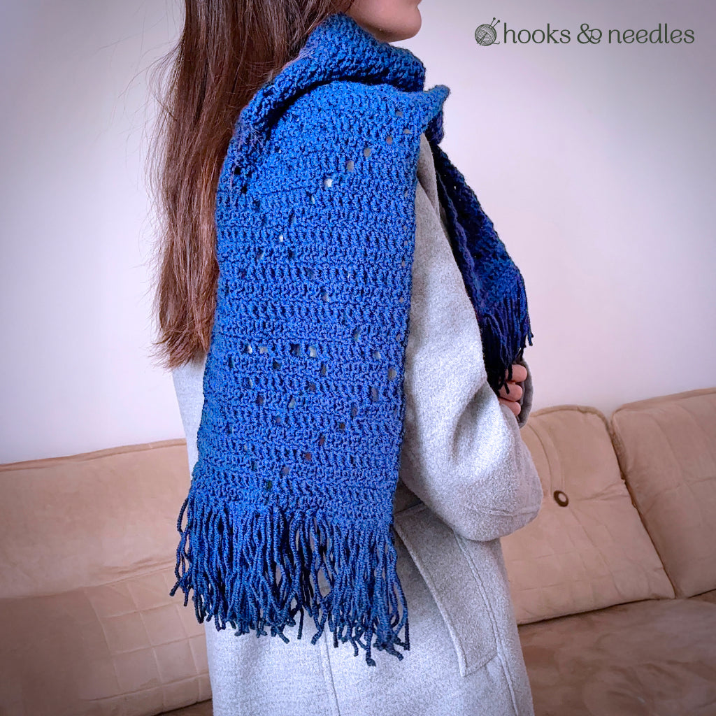 A person in a gray coat showcases the Classic Argyle Scarf, made with a beginner-friendly crochet pattern, featuring blue yarn and fringe, while standing before a beige couch.