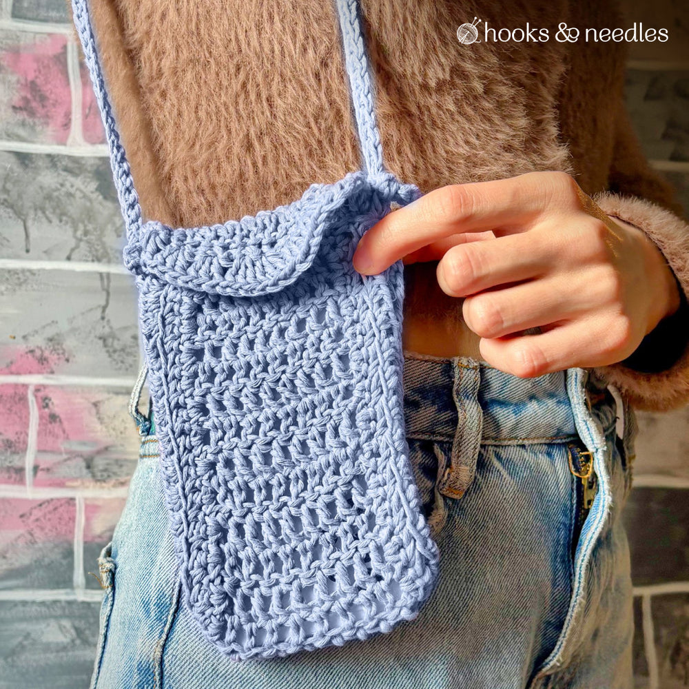 A person in jeans and a brown sweater holds a light blue Phone Pouch Crochet Pattern with a customizable strap, standing before a gray and pink wall.