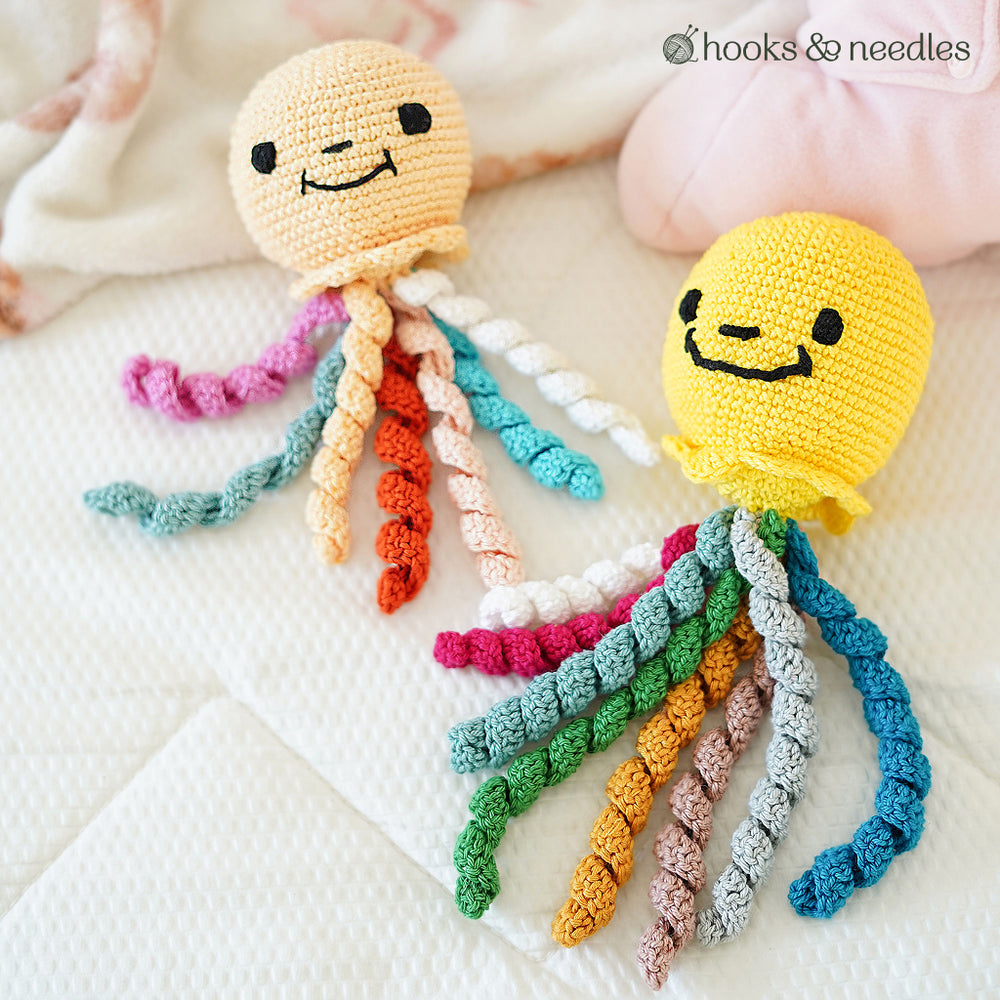 Two beginner-friendly Octopus Toy Crochet Pattern designs with smiley faces and colorful spiral tentacles rest on a white quilt, highlighting the charm of handmade crochet.