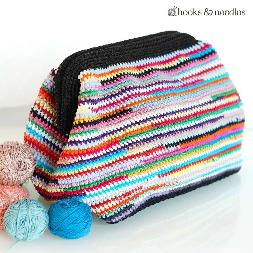 An Upcycled Color Clutch, made from yarn scraps using the Upcycled Color Clutch Crochet Pattern, rests on a white surface beside three yarn balls in pink, blue, and peach. "Hooks & needles" appears in the top right corner.
