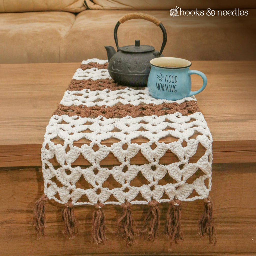 The Table Runner Crochet Pattern features a handcrafted design in brown and white, displayed on a wooden table with a black teapot and a blue "GOOD MORNING" mug.