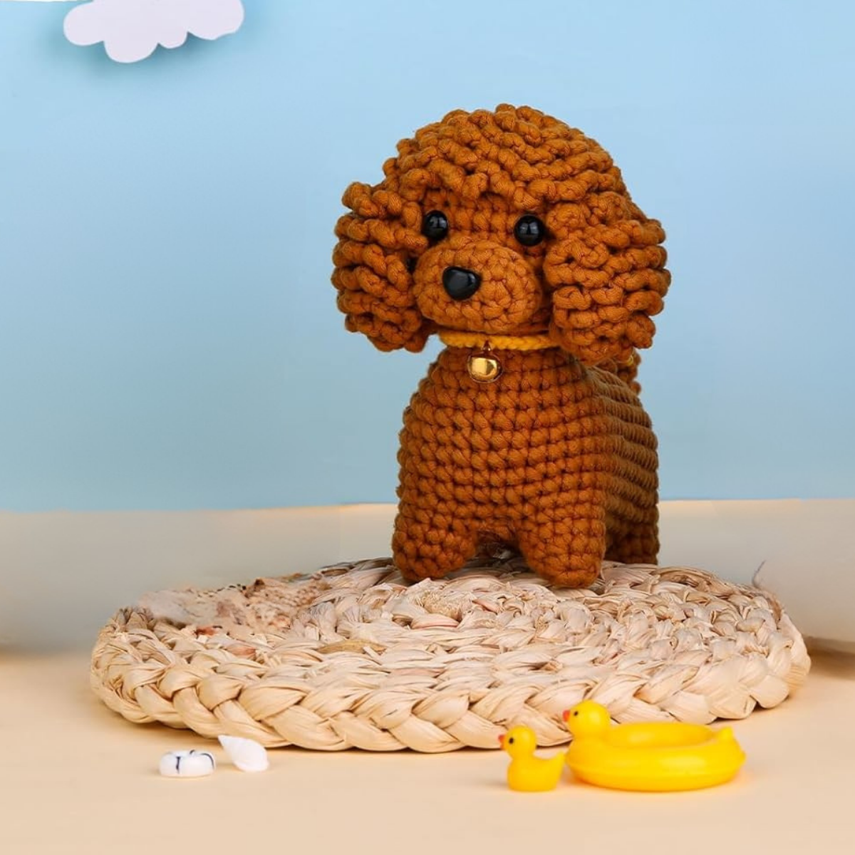 The Crochet Poodle Kit features a handmade poodle plush on a woven mat with a toy duck and yellow ducklings, set against a blue background—ideal for beginners seeking an easy crochet project.