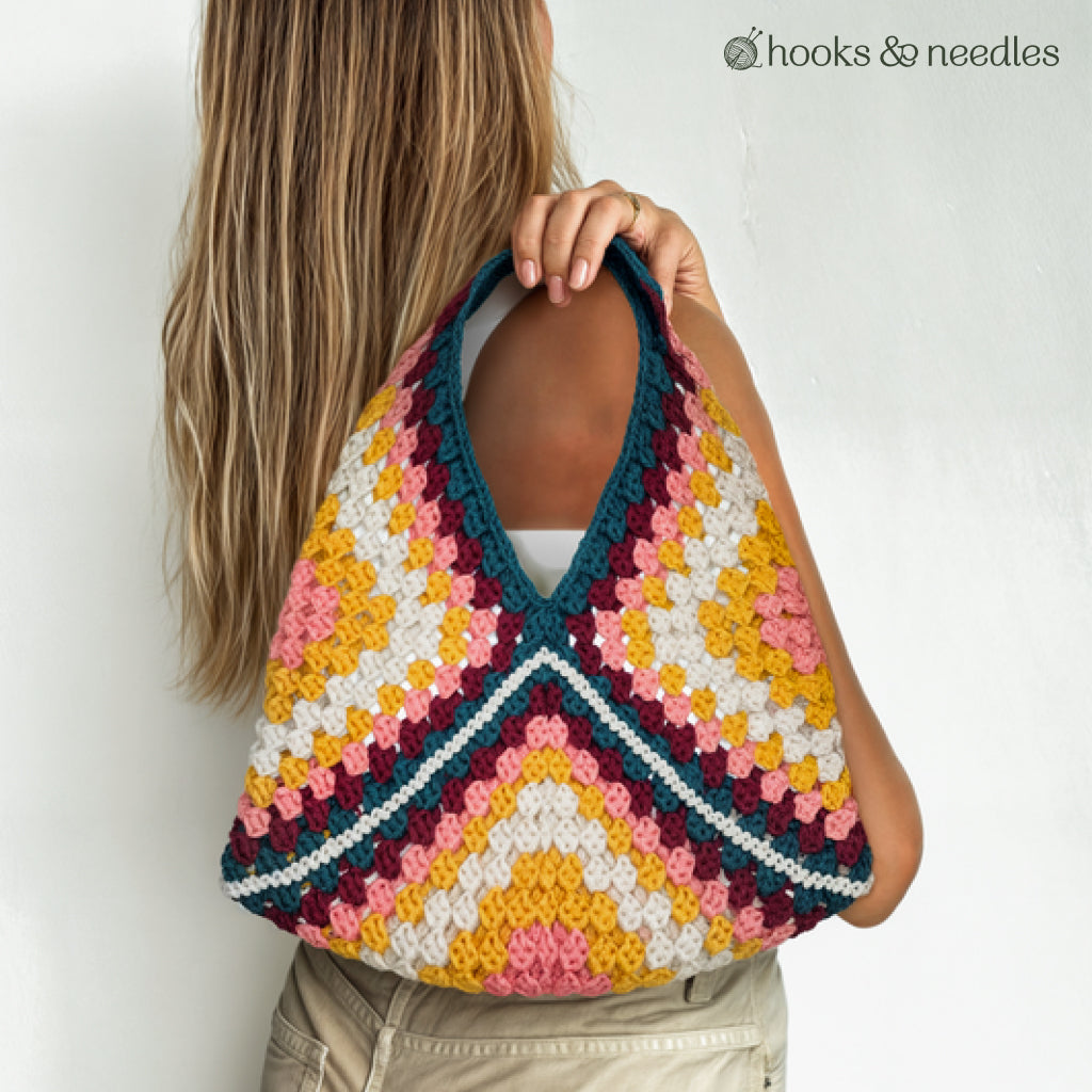 A person with long blonde hair holds a large Granny Square Tote Bag Crochet Pattern, showcasing vibrant geometric crochet designs in yellow, pink, teal, and white.