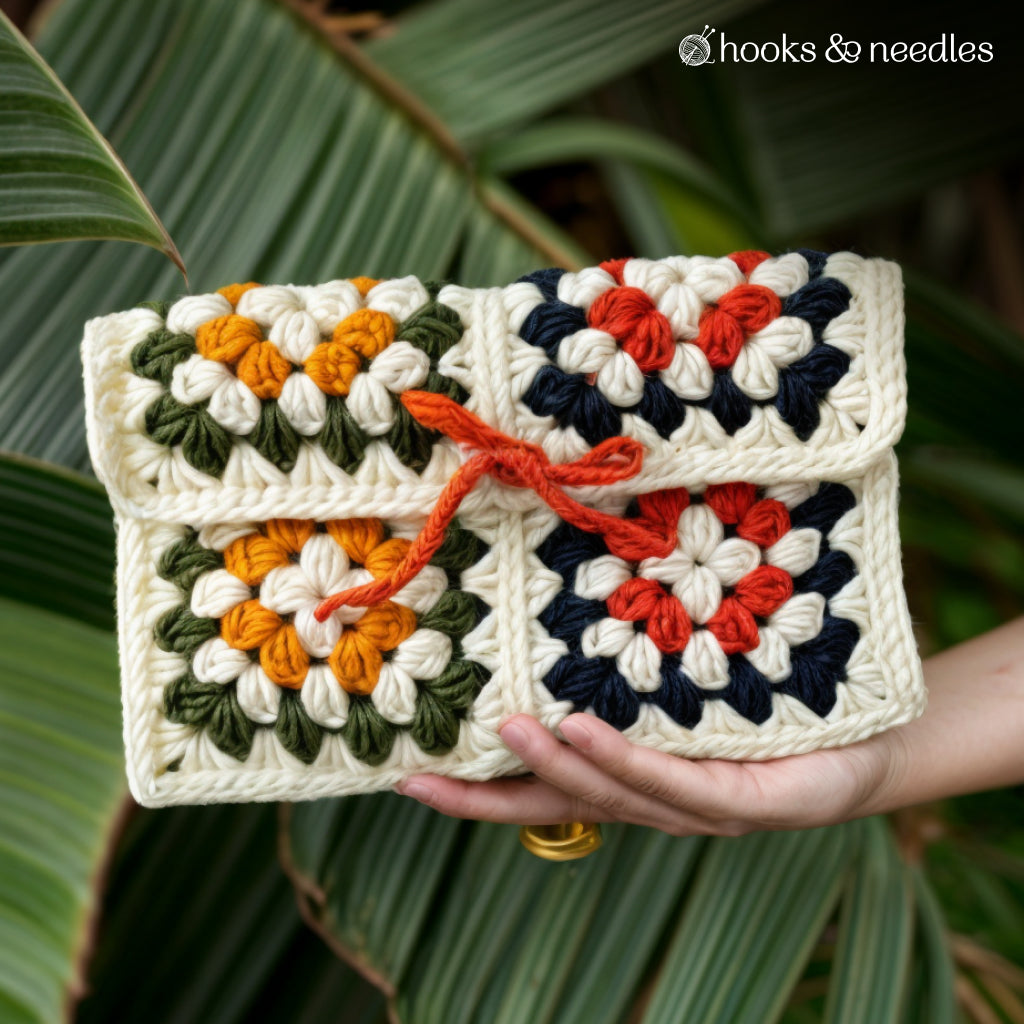 A hand holds the Cleo Clutch, made from the Cleo Clutch Crochet Pattern, featuring vibrant granny squares and a red yarn tie at the front. Green leaves are in the background.