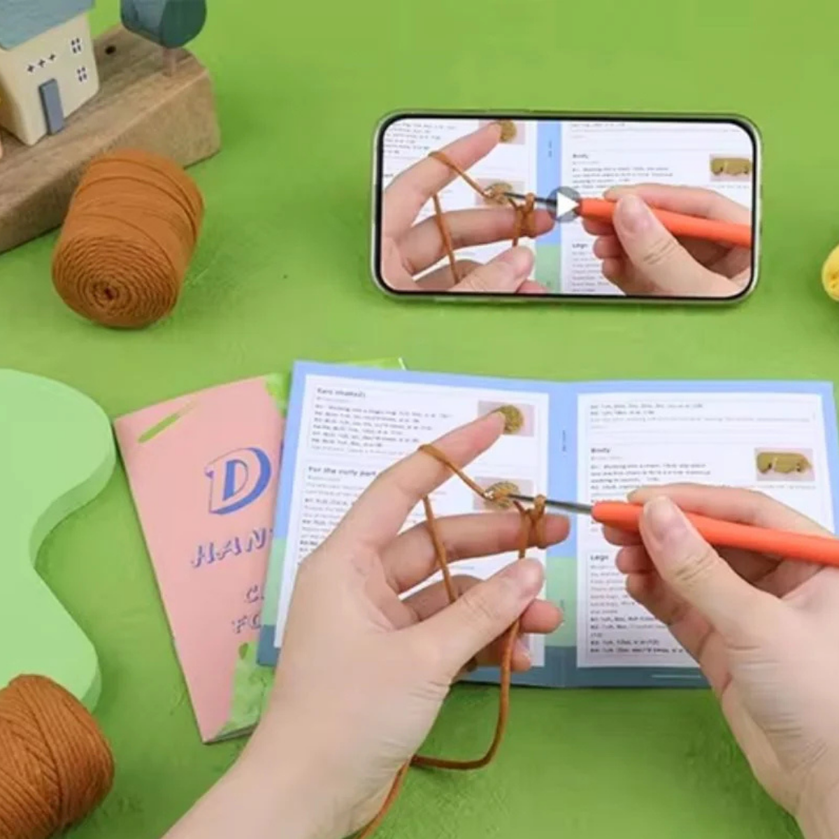 Using the Crochet Poodle Kit and a phone tutorial, a person prepares to create a handmade poodle plush, holding yarn and hook at a green table with crafting supplies.