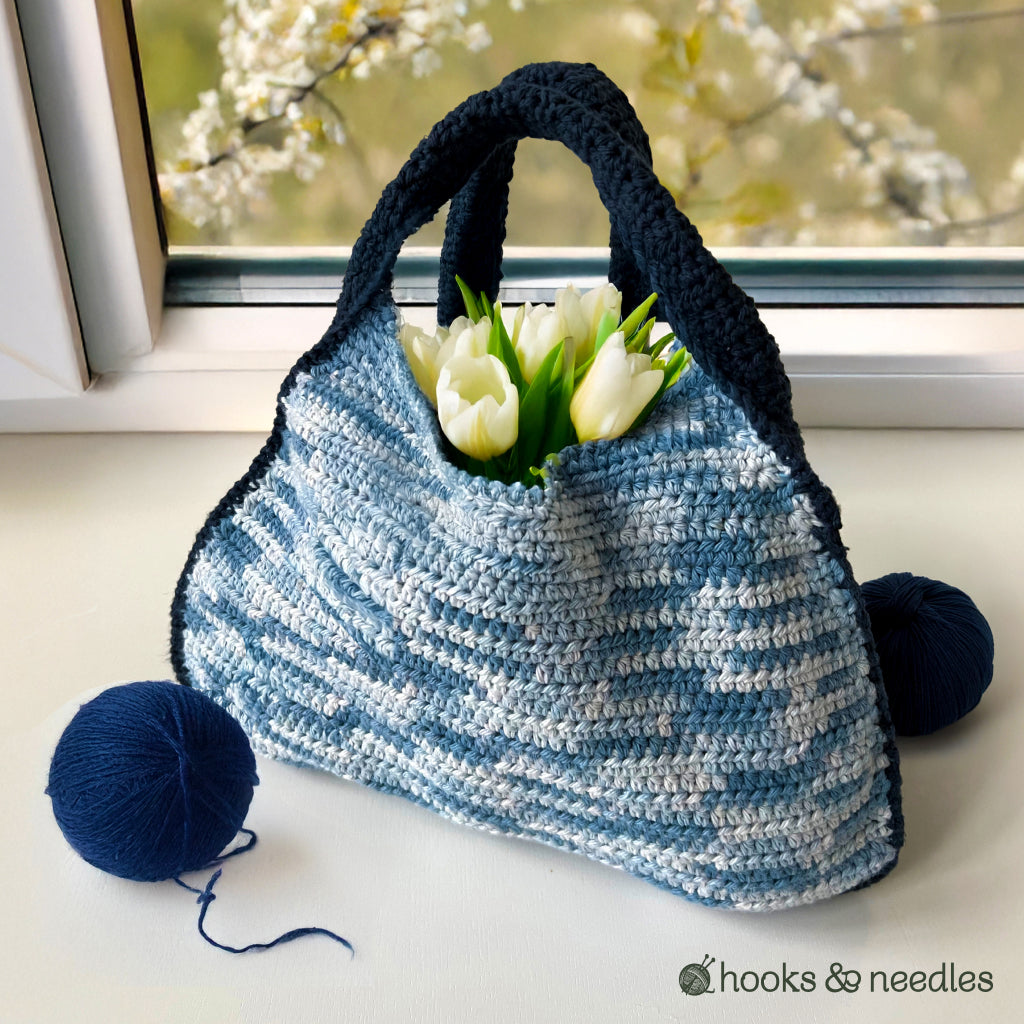 A Two-Tone Bag made from the Crochet Pattern features blue and white colors with black handles, holding white tulips. Nearby are blue yarn balls, creating a stylish windowsill display perfect for intermediate crocheters.