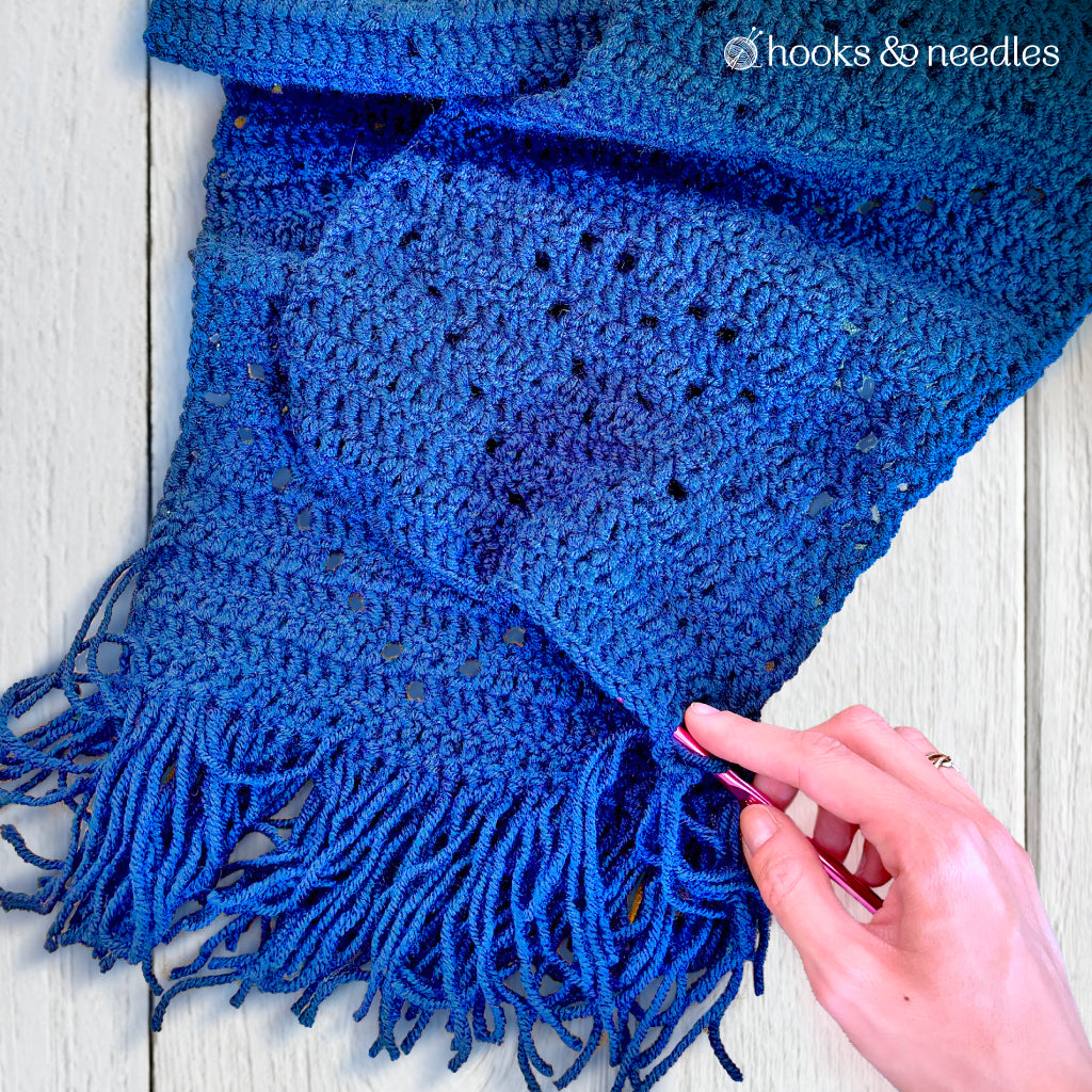 A hand holds the fringe of a blue scarf on a light wooden surface, displaying the Classic Argyle Scarf Crochet Pattern as a stylish accessory, with "hooks & needles" text in the corner.