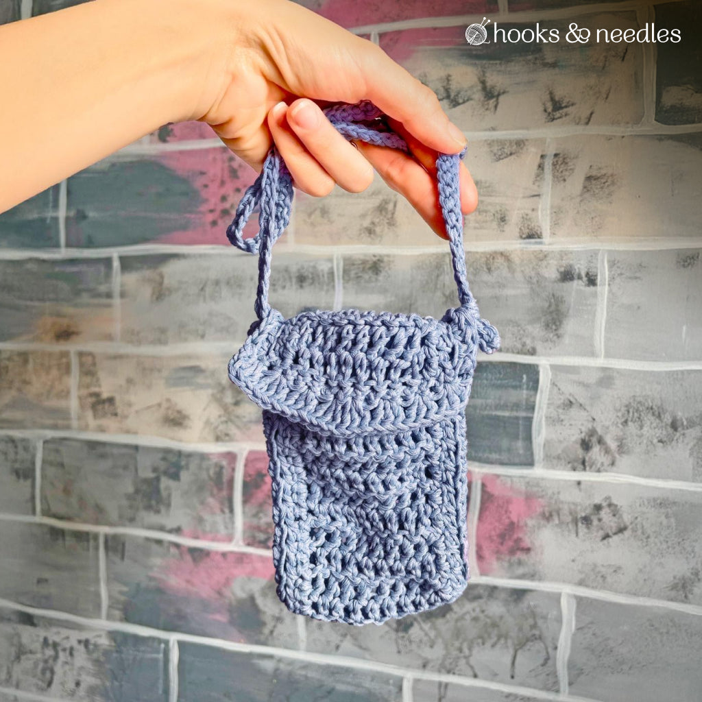 A hand displays a small, blue pouch made from the Phone Pouch Crochet Pattern, featuring a flap and customizable strap, in front of a painted brick wall.