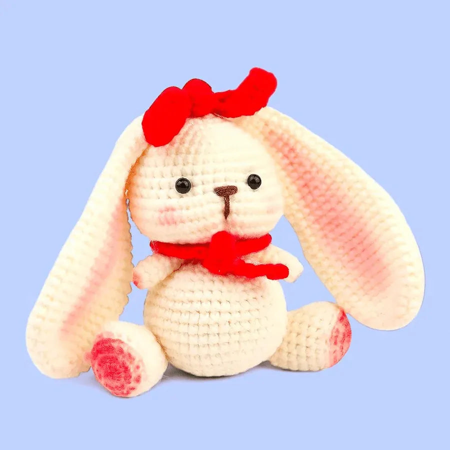 A plush white bunny with long ears, pink accents, a red bow, and scarf—handcrafted from the Crochet Rabbit Kit—sits against a light blue background.
