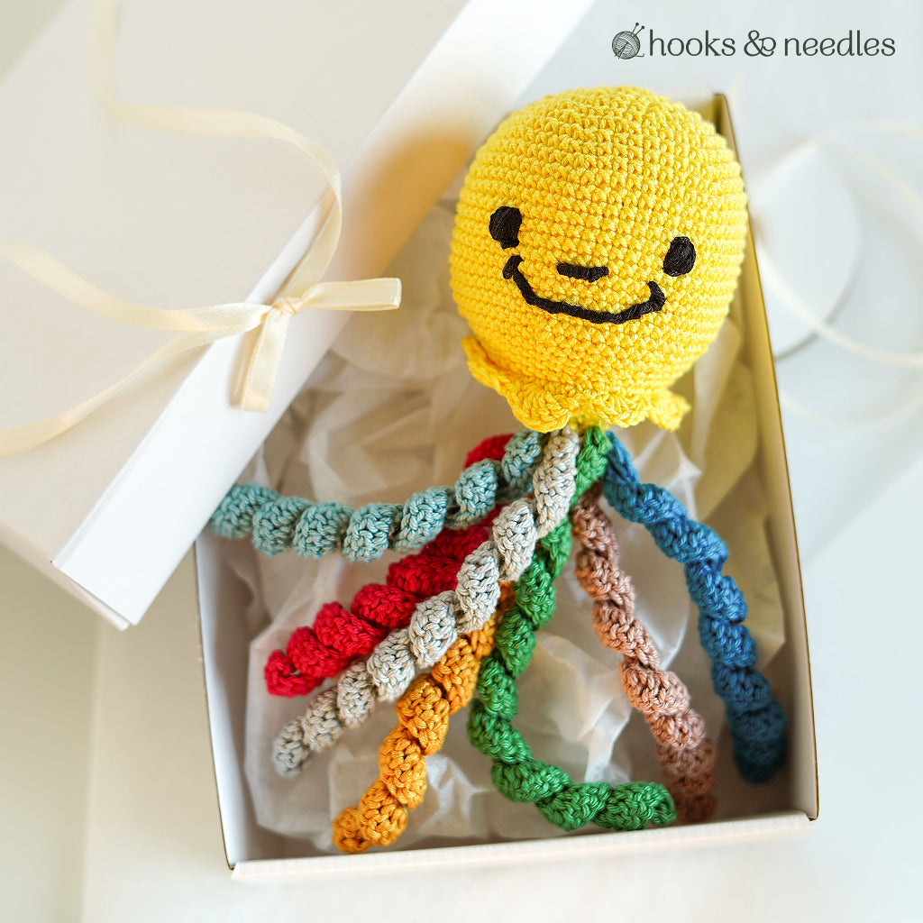 An Octopus Toy Crochet Pattern features a cheerful yellow octopus with a smiling face and multicolored tentacles, displayed in a tissue-lined gift box.