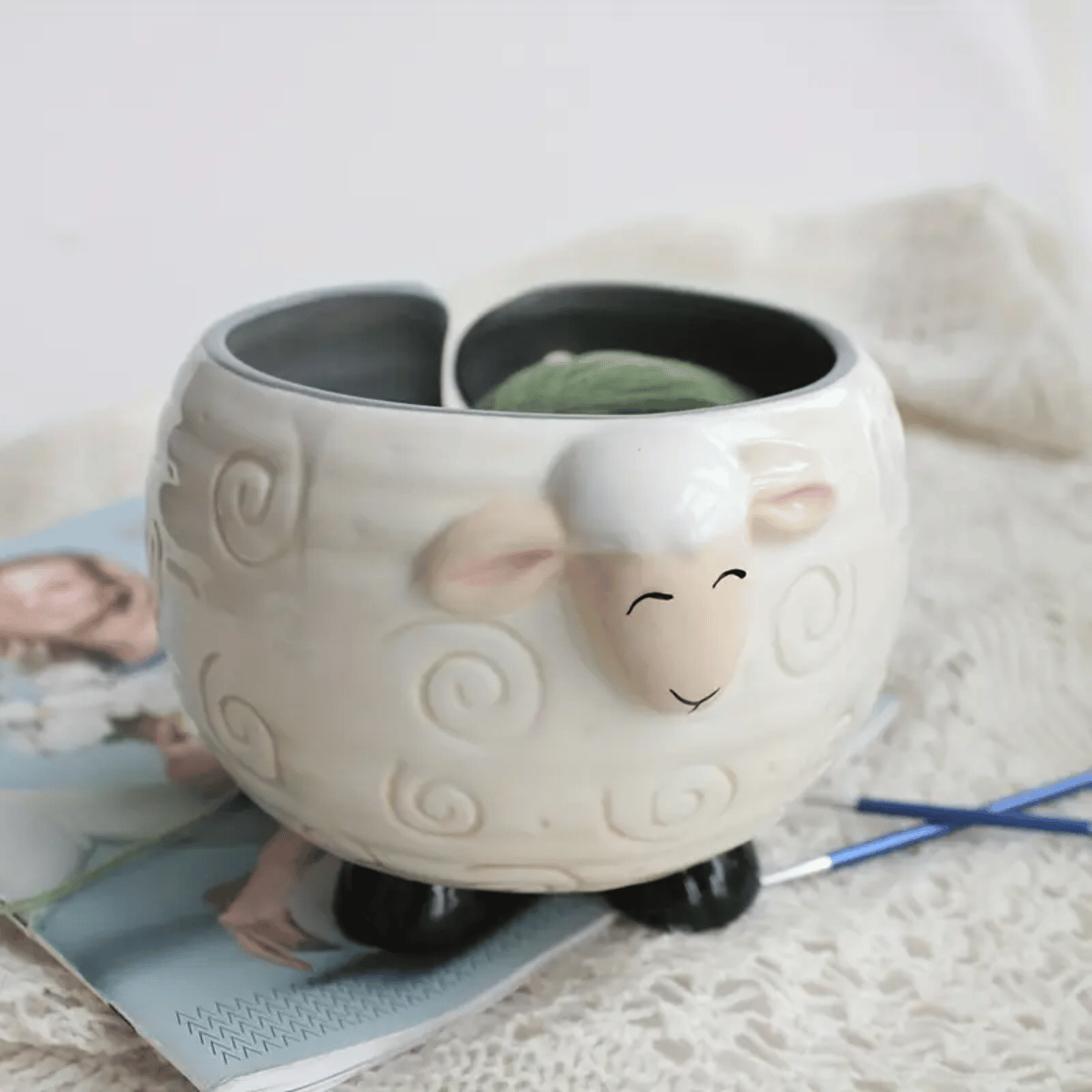 The Sheep Ceramic Yarn Bowl, shaped like a cute sheep, holds a green yarn ball and rests on knitted fabric with a magazine and knitting needles—an adorable crochet accessory for any maker.