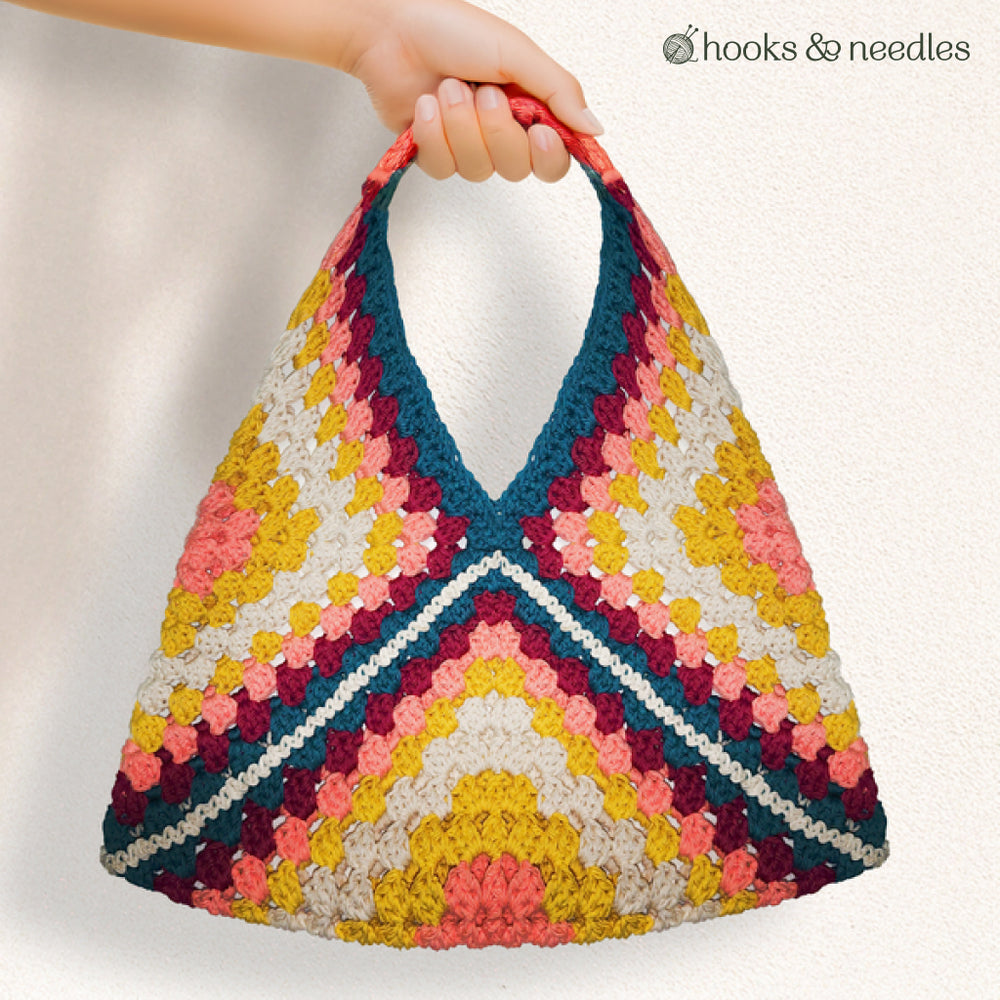 A hand displays the Granny Square Tote Bag Crochet Pattern featuring a vibrant geometric crochet design. The plain background highlights its beginner-friendly style, with the "hooks & needles" logo in the top right corner.