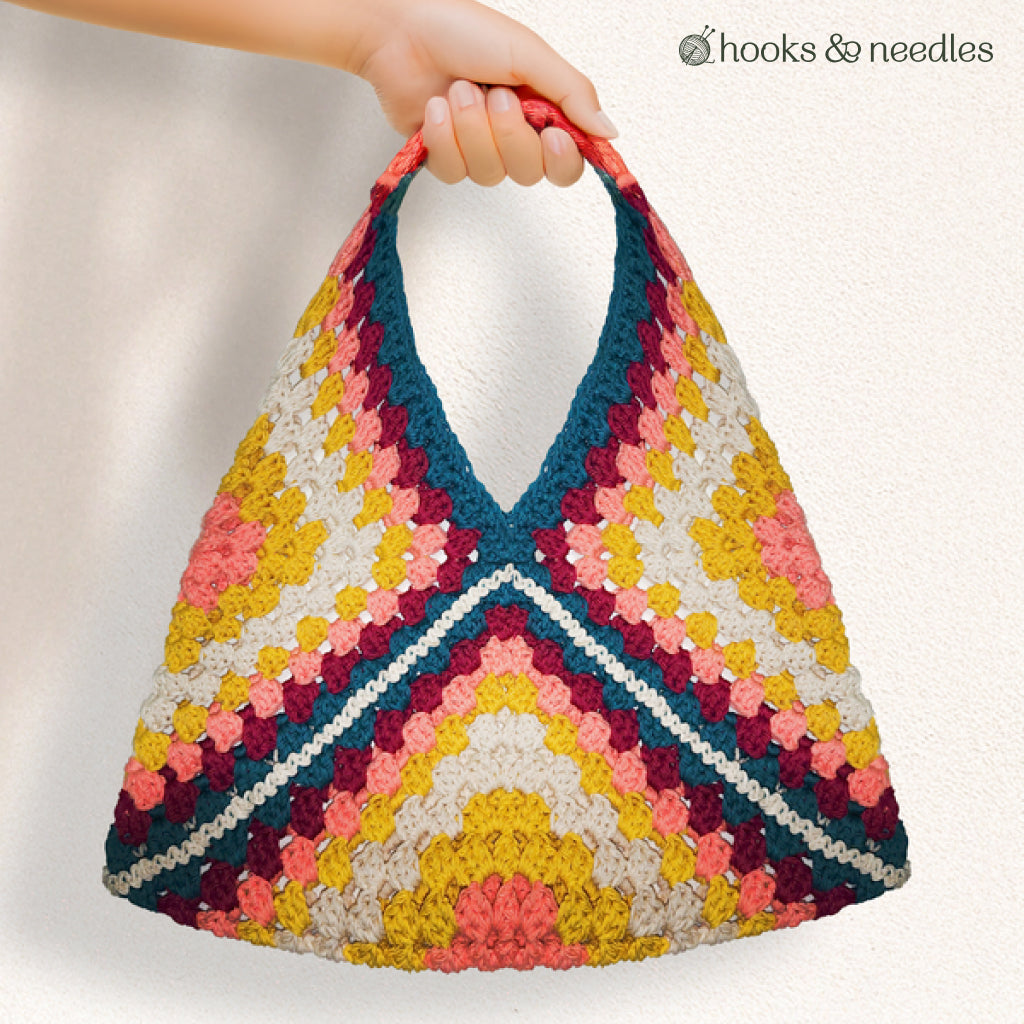 A hand displays the Granny Square Tote Bag Crochet Pattern featuring a vibrant geometric crochet design. The plain background highlights its beginner-friendly style, with the "hooks & needles" logo in the top right corner.