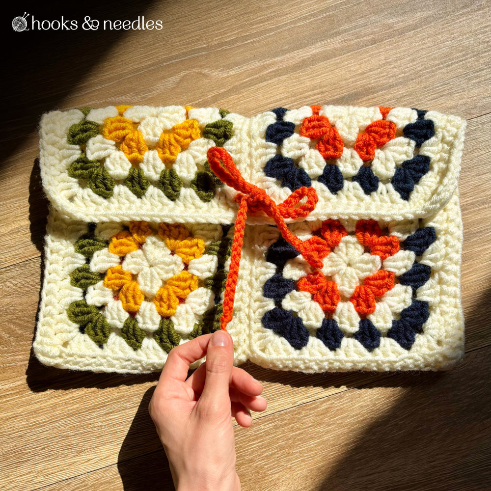 A hand displays the Cleo Clutch Crochet Pattern—a pouch featuring classic granny squares in yellow, green, orange, navy, and cream yarn with an orange tie—ideal for crochet enthusiasts and shown on a wooden surface.