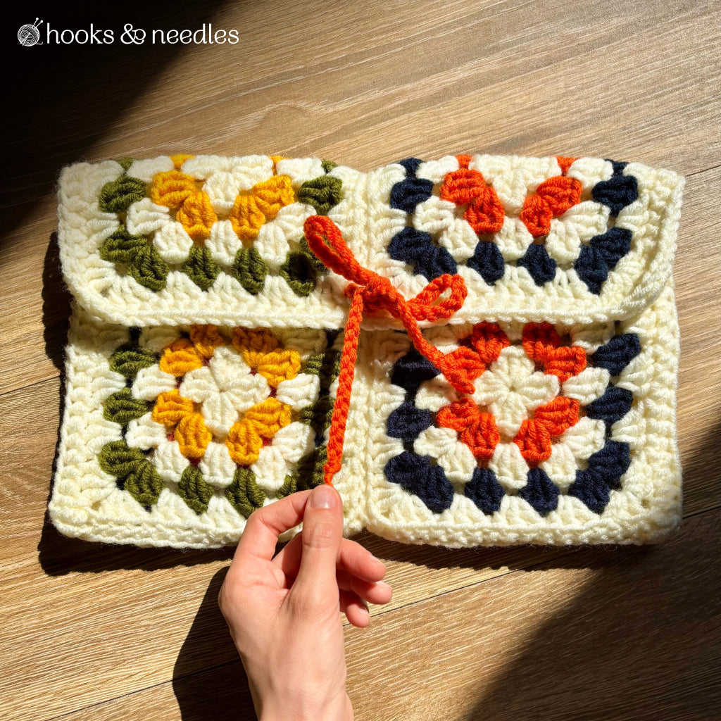 A hand displays the Cleo Clutch Crochet Pattern—a pouch featuring classic granny squares in yellow, green, orange, navy, and cream yarn with an orange tie—ideal for crochet enthusiasts and shown on a wooden surface.