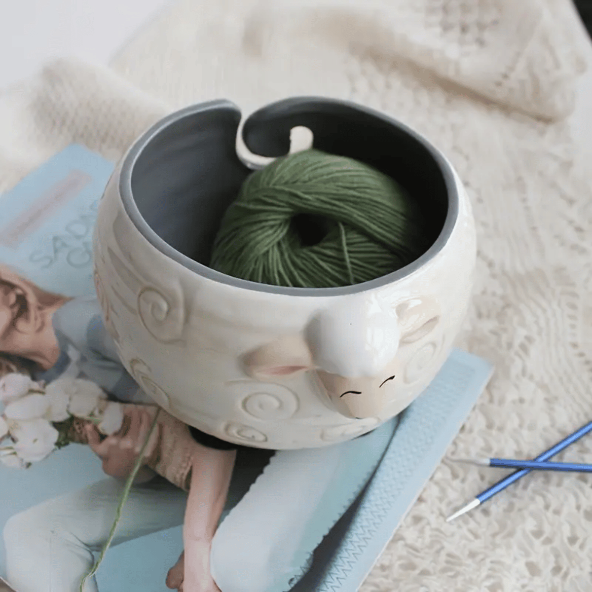 The Sheep Ceramic Yarn Bowl, an ideal crochet accessory, holds green yarn. It sits on a magazine and cream knitted fabric with knitting needles nearby.