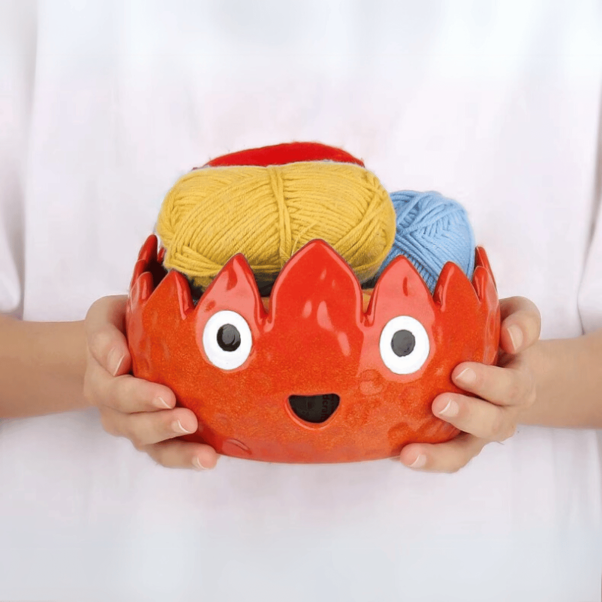 A person holds the Flame Ceramic Yarn Bowl, a red ceramic bowl with eyes and a mouth, containing yellow and blue yarn balls.