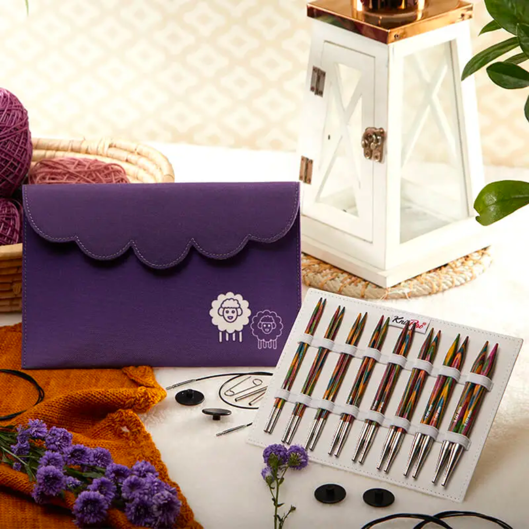 A purple clutch with sheep designs on a table alongside knitting supplies and the Deluxe Wooden Knitting Needle Set.