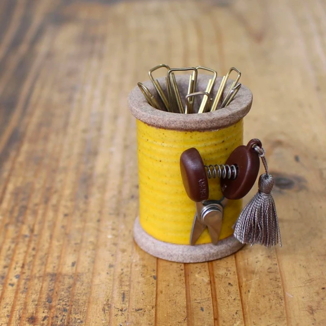 A Small Scissors keychain, equipped with precision mini sewing scissors and a tassel, accompanies gold paperclips on a yellow ceramic spool, all arranged on a wooden surface. Compact and portable, this stylish ensemble enhances desk functionality.