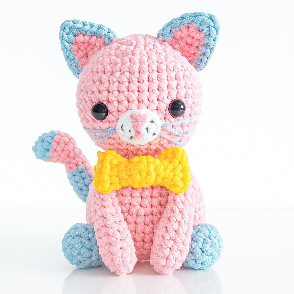 Luna The Cat is a crocheted amigurumi plush toy in pink and blue with black button eyes and a yellow bowtie, sitting upright against a white background—ideal for intermediate crochet enthusiasts.