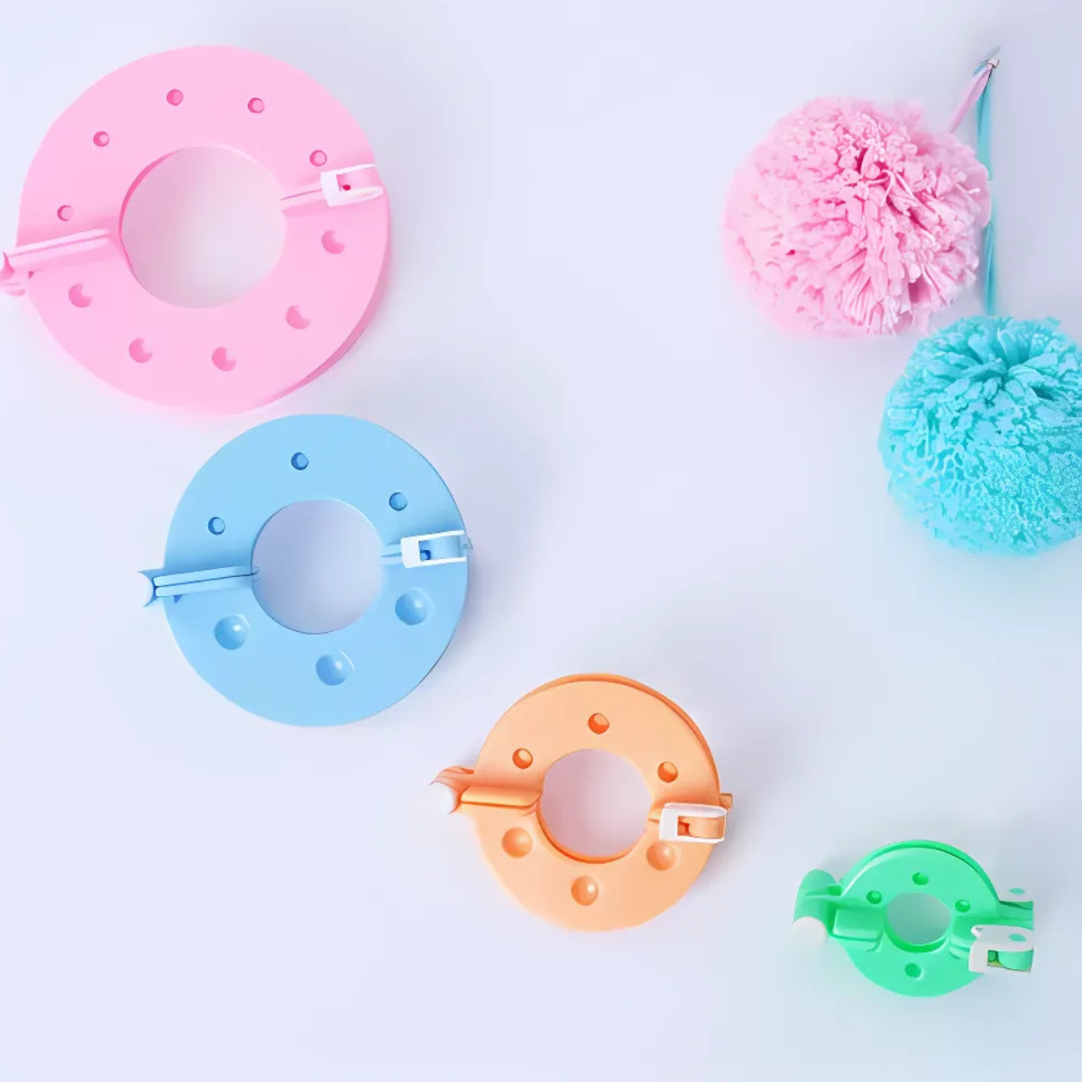 The Pom Pom Maker Set includes four plastic tools in pink, blue, orange, and green on a white surface, with two finished yarn pom poms nearby—ideal for crafting colorful decorations.
