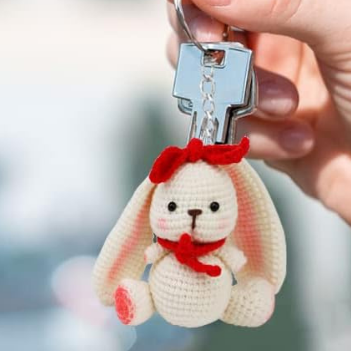 A hand holds a keychain made with the Crochet Rabbit Kit, featuring a crocheted white bunny with long ears, a red bow on its head, and a red scarf around its neck.