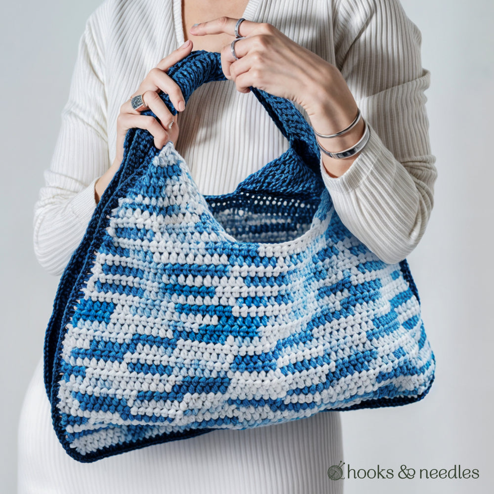 A person in a white ribbed top holds the "Two-Tone Bag Crochet Pattern"—a large blue and white crocheted bag with a thick handle, ideal for intermediate crocheters. The "hooks & needles" logo appears in the corner.
