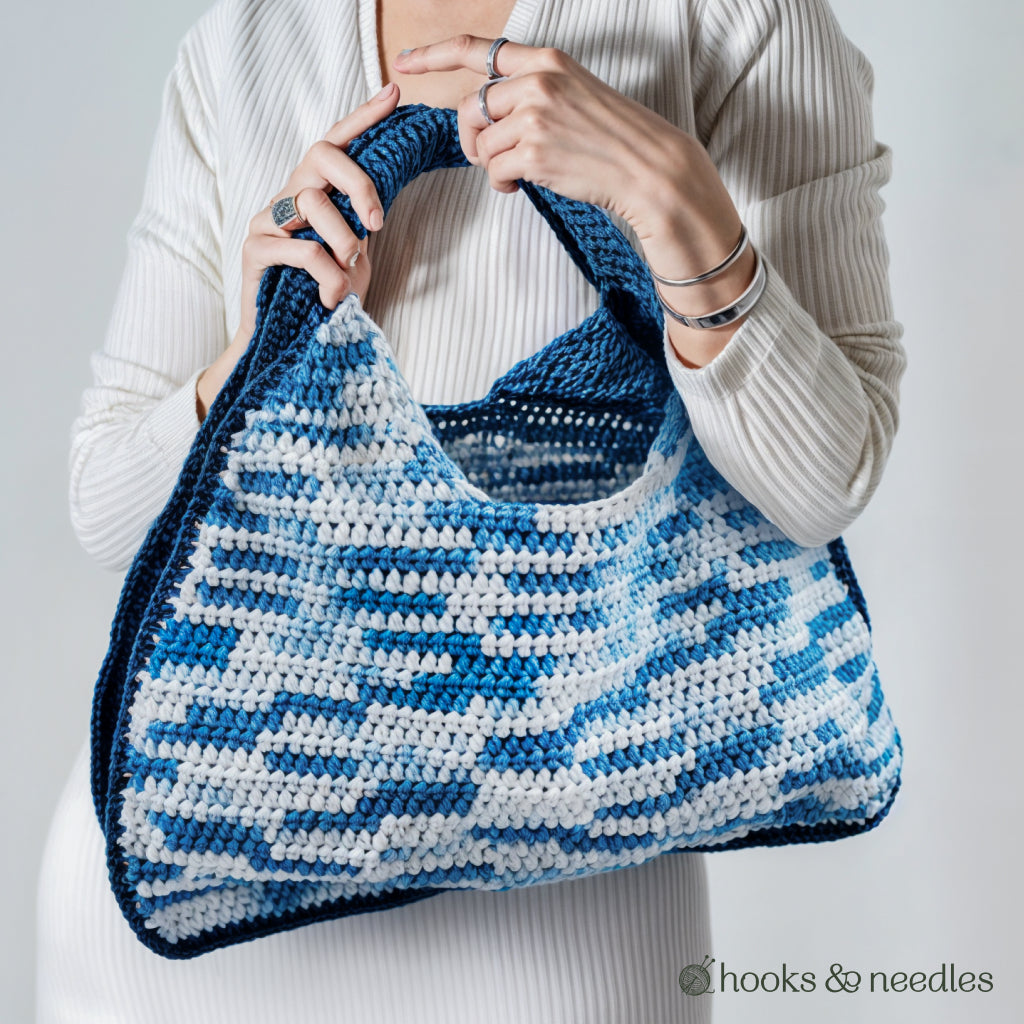 A person in a white ribbed top holds the "Two-Tone Bag Crochet Pattern"—a large blue and white crocheted bag with a thick handle, ideal for intermediate crocheters. The "hooks & needles" logo appears in the corner.