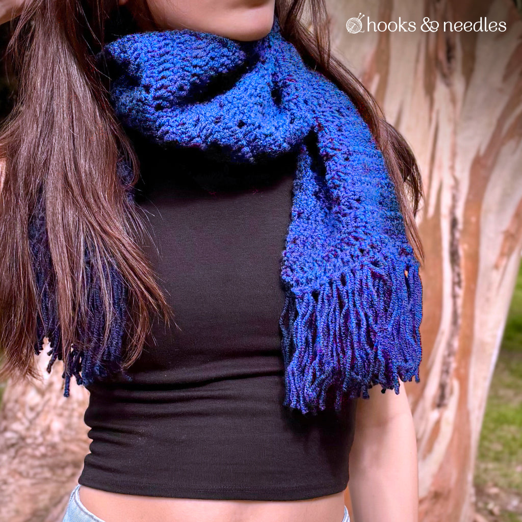 A person wearing a blue Classic Argyle Scarf Crochet Pattern with fringes over a black crop top, standing outdoors near a tree.
