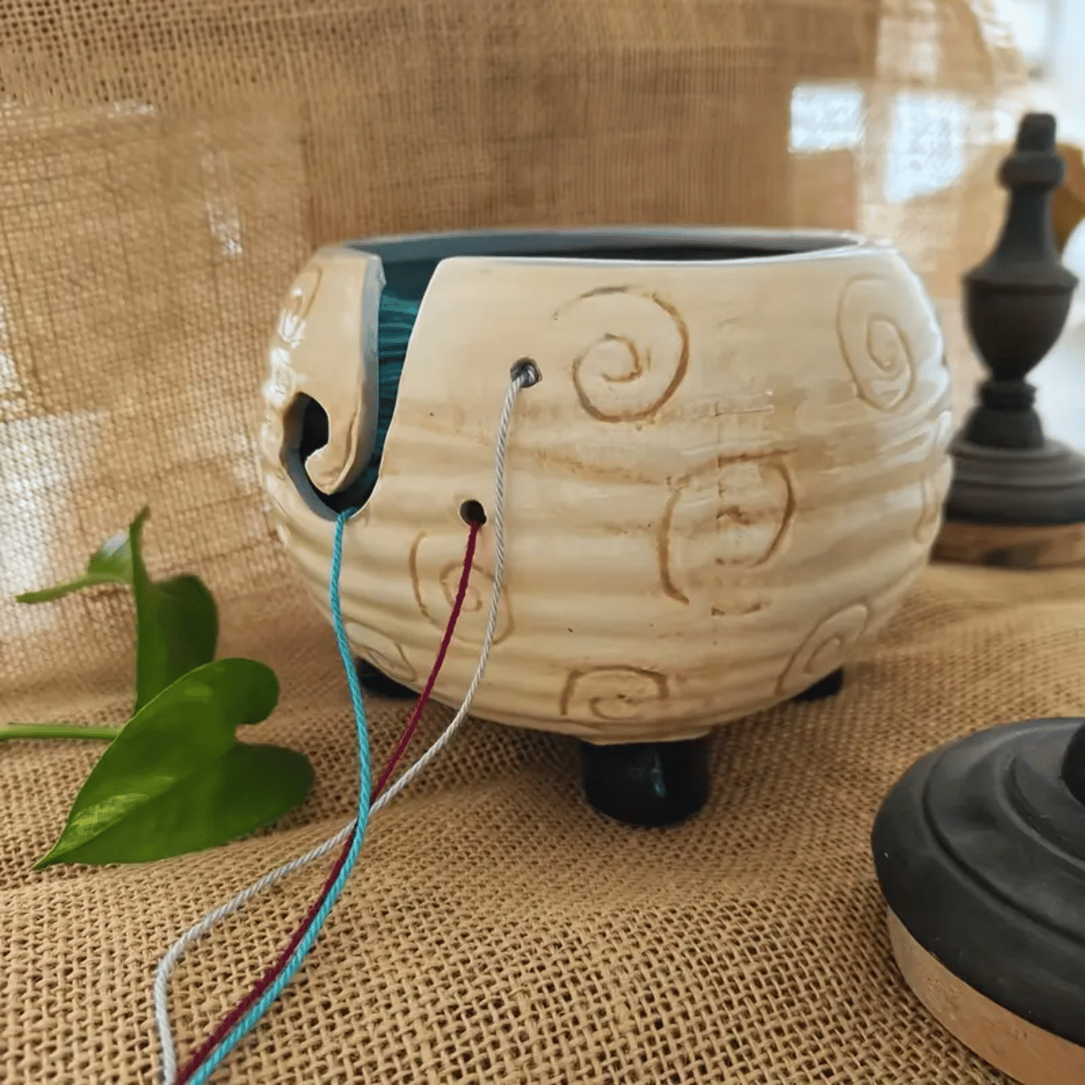The Sheep Ceramic Yarn Bowl, crafted from ceramic and designed to hold three strands of yarn, features a charming sheep motif. In the background, a green plant leaf and blurred chess pieces rest on burlap.