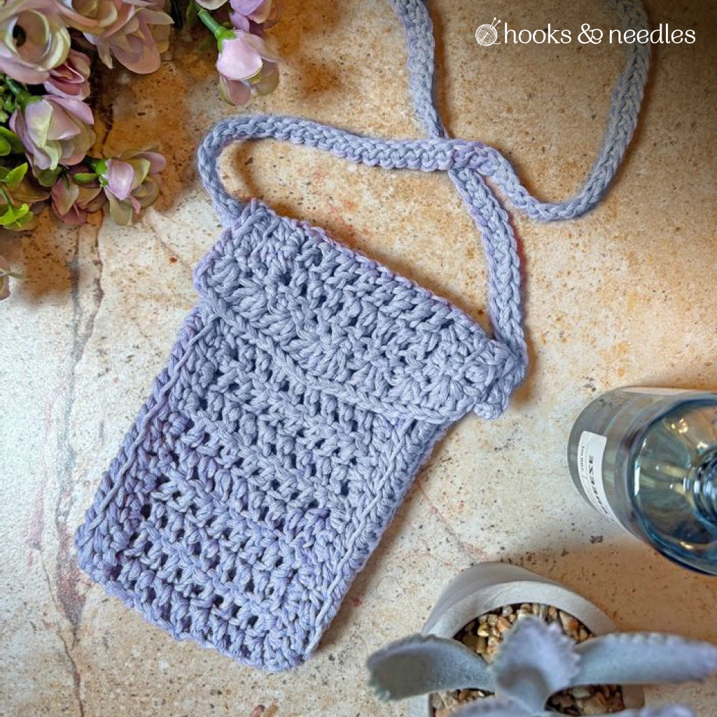The Phone Pouch Crochet Pattern is shown as a small lavender crochet bag with a long, adjustable strap on a stone surface near plants and a water bottle—ideal for carrying your phone or accessories.