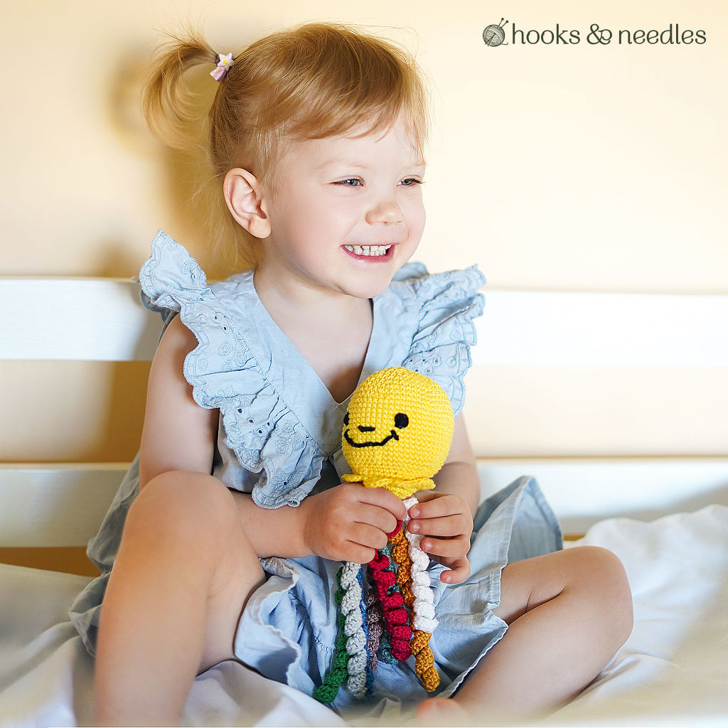 A young child with blonde pigtails smiles on a bed, holding a yellow octopus toy with multicolored tentacles made using the Octopus Toy Crochet Pattern.
