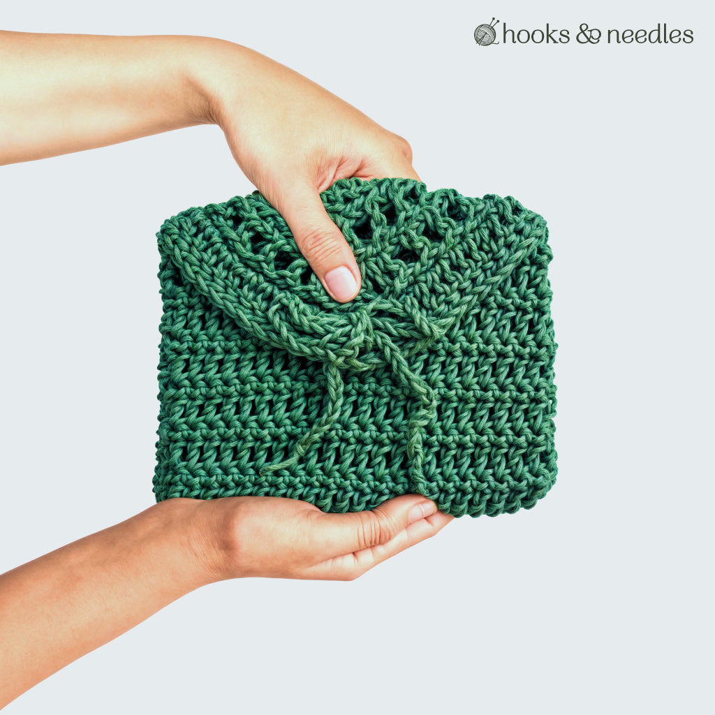 Two hands hold a green Petite Purse Crochet Pattern pouch with a drawstring—perfect for daily essentials. The "hooks & needles" logo appears in the upper right. This beginner-friendly pattern is both stylish and practical.