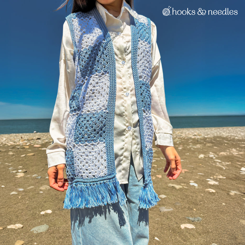 A person stands on a beach in a white shirt, blue jeans, and a stylish vest made using the Checkered Vest Crochet Pattern. The clear sky and sea in the background highlight the unique checkered crochet design with fringe.