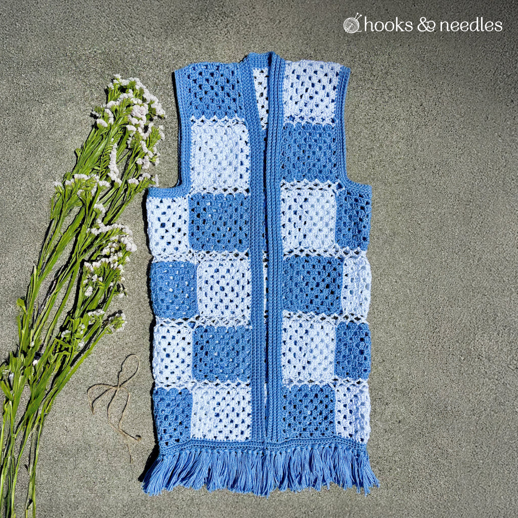 The Checkered Vest Crochet Pattern features blue and white granny squares with playful fringe, displayed on a gray background next to a sprig of white flowers.