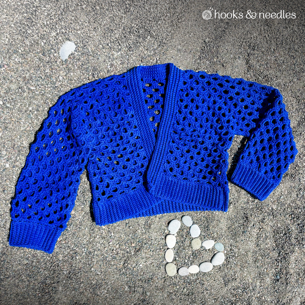 The Honeycomb Stitch Cardigan Crochet Pattern features a royal blue, lacy, openwork design—shown laid flat on sand beside a heart of white stones—ideal inspiration for advanced crocheters.