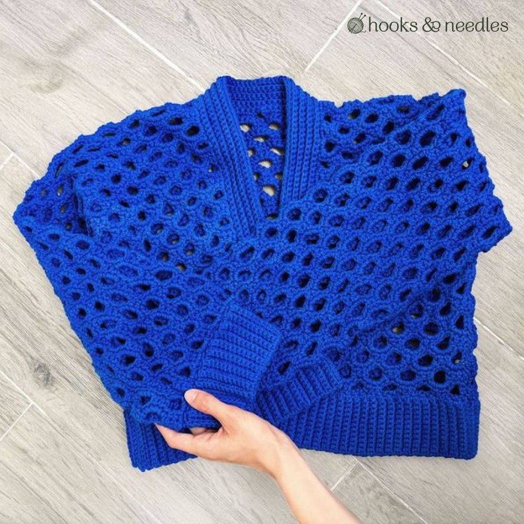 A hand holds the Honeycomb Stitch Cardigan, crocheted with a blue openwork mesh pattern, neatly folded on a light wood floor.