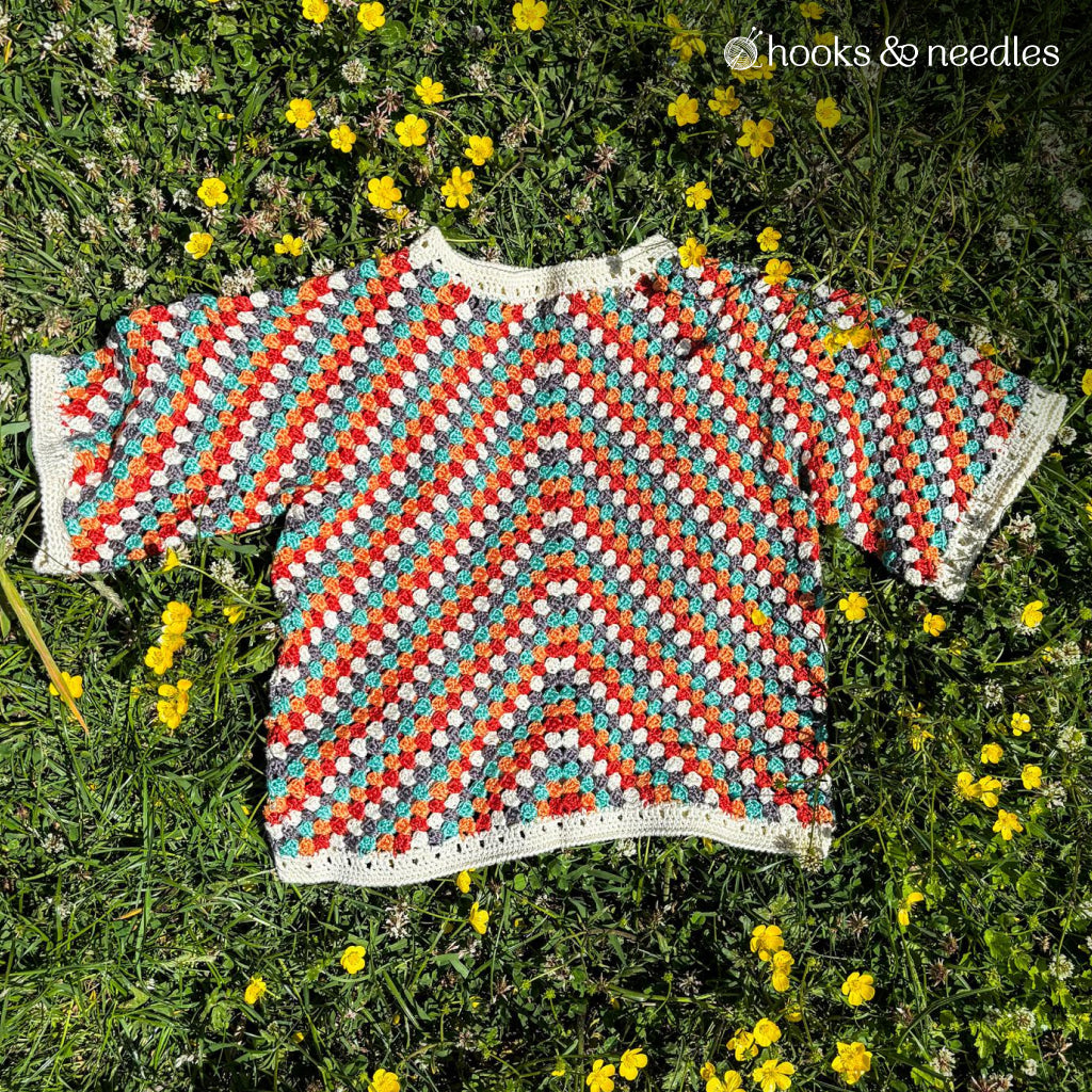 The Sienna Arrow Sweater Crochet Pattern features a crocheted short-sleeve top with red, blue, and white stripes in soft bamboo yarn, shown laid flat on grass adorned with yellow flowers.