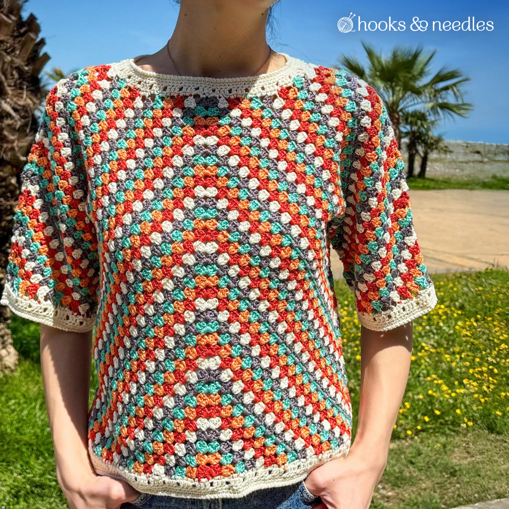 Someone stands outside near grass and palm trees, wearing a colorful short-sleeve top with a chevron design, handmade from the Sienna Arrow Sweater Crochet Pattern.