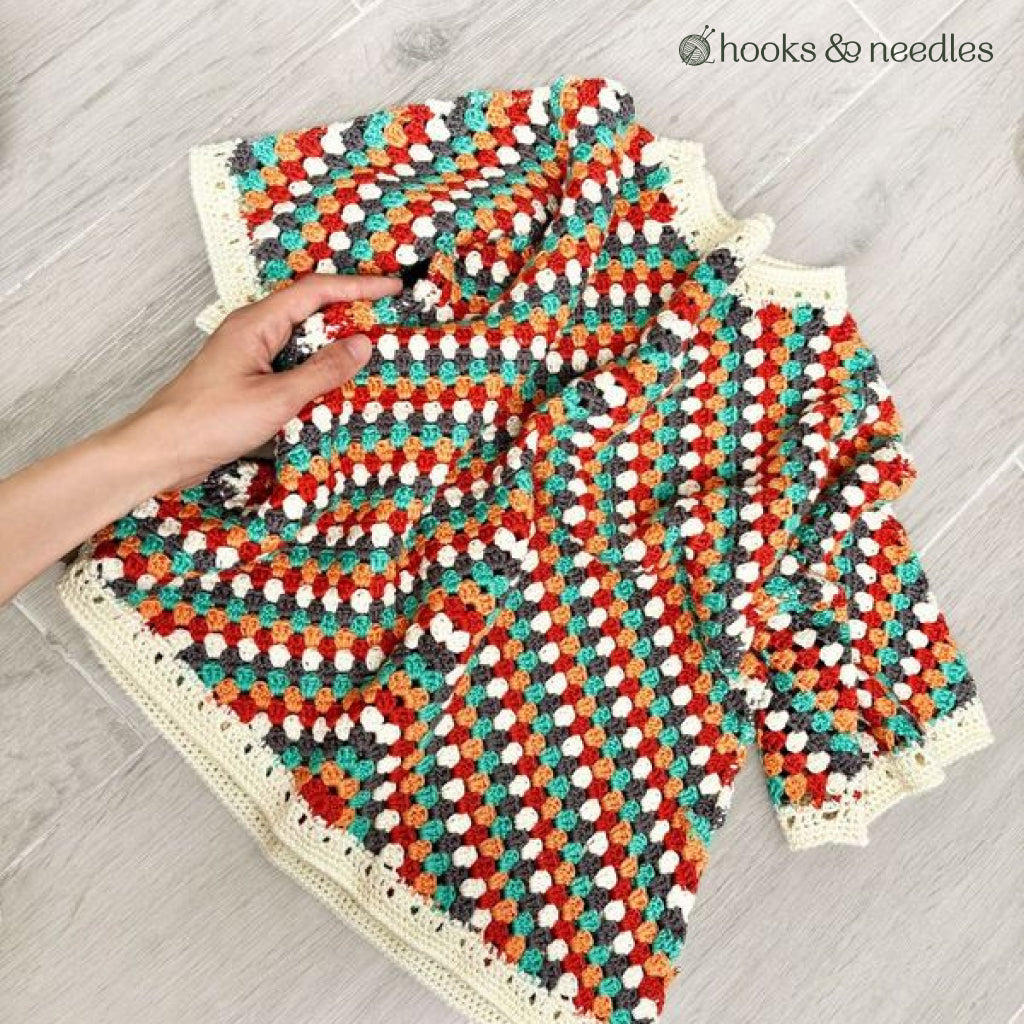 A hand holds the Sienna Arrow Sweater Crochet Pattern—a colorful, crocheted cardigan with cream borders, made from soft bamboo yarn and laid flat on a light wood surface.