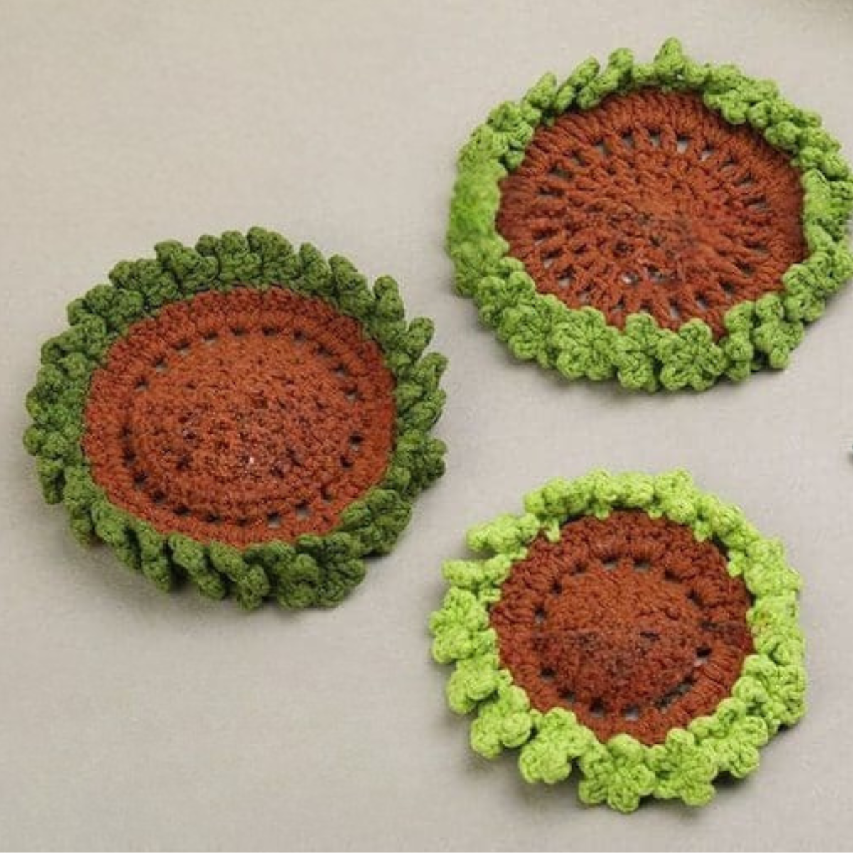 The Plant Crochet Coaster Kit features three round handmade wool coasters with brown centers and green textured edges, evoking sunflowers or plants, displayed on a neutral background.