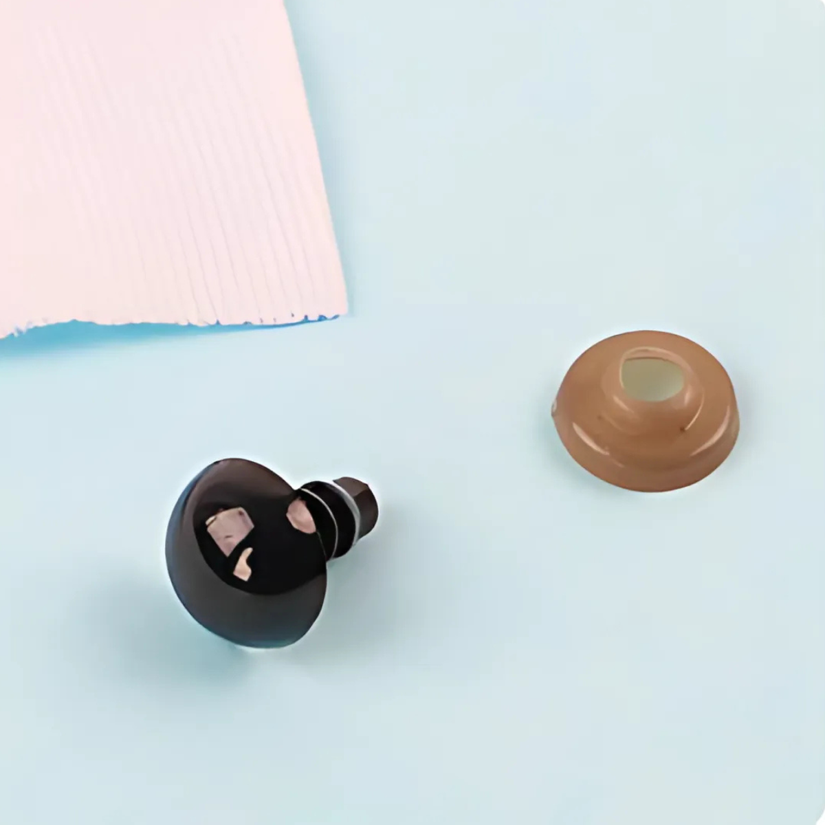 A black wireless earbud and a beige silicone ear tip rest on a light blue surface with torn white paper, resembling tiny accessories like Safety Eyes for DIY, often used in amigurumi or crochet projects.