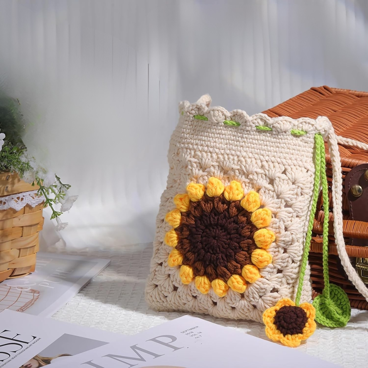 The Sunflower Crochet Bag Kit, featuring a cream base and large yellow sunflower design, is shown next to a wicker basket and magazine on a white textured surface.