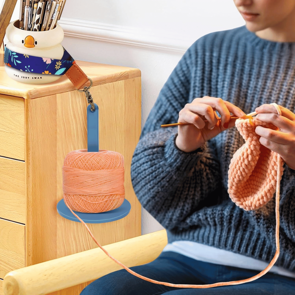 A person uses a Wrist Yarn Holder to crochet hands-free with peach yarn, wearing a blue sweater, and the yarn is secured to a wooden drawer.
