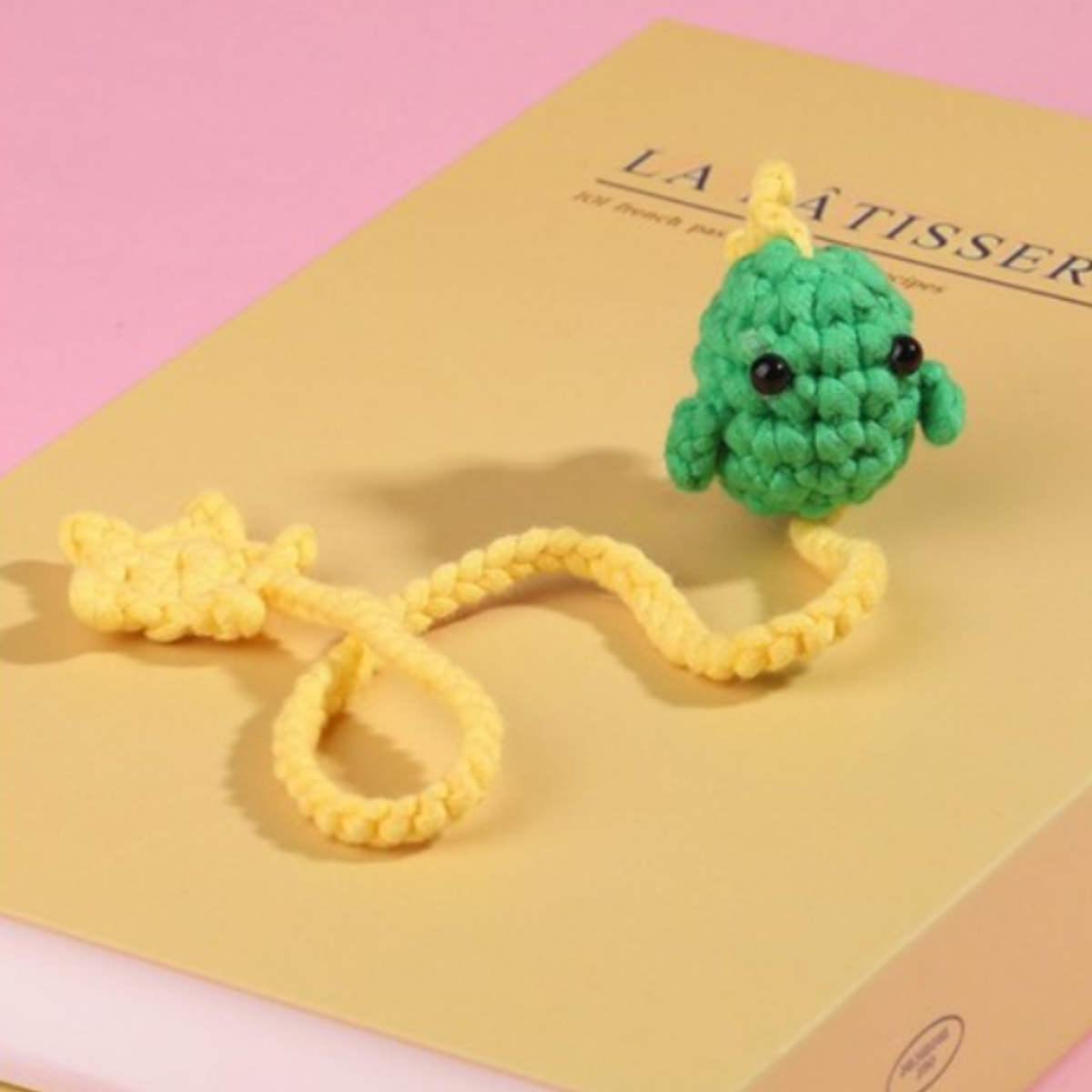 A green chameleon with a yellow tail and feet, made from the Cute Animal Crochet Bookmark Kit, sits on a pale yellow book against a pink background.